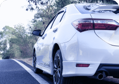 A white car from Promex Auto is parked on the side of a road surrounded by greenery. The car's rear and part of its side are visible, showcasing a rear spoiler and sporty design. The road curves slightly, leading into a wooded area filled with trees and bushes.