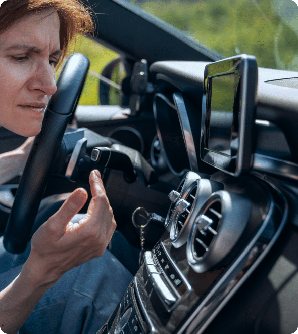 A person sitting in a car looks frustrated as they hold a smartphone in one hand while gripping the steering wheel with the other. The car's dashboard, including an infotainment screen and various controls, is visible. The person appears to be searching for directions.