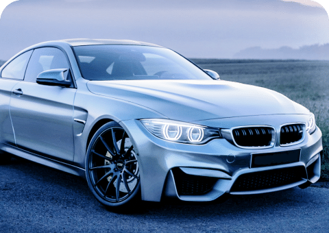 A sleek silver sports car, possibly a BMW model from Promex Auto, is parked on a paved area during twilight. The car's headlights are on, and the background features a blurred view of a grassy field and a hazy sky.