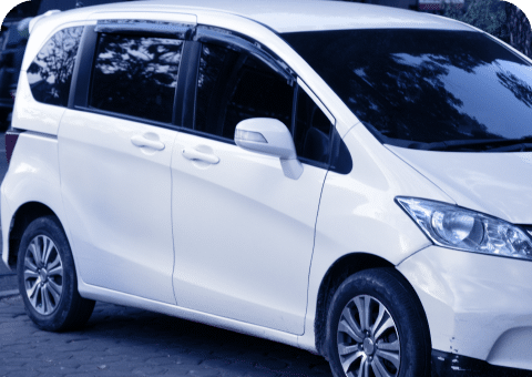 A white minivan from Promex Auto is parked on a paved surface. The vehicle has tinted windows and the surroundings reflect trees and foliage, indicating it is likely parked outdoors near a wooded area. The image shows the front and side profile of the minivan, possibly awaiting auto repair services.