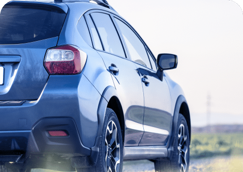 A blue crossover SUV from Promex Auto is parked outdoors on a sunny day, with a blurred background of grass and distant mountains. The shot is angled from the rear side of the vehicle, highlighting the car's rear lights, window reflections, and metallic paint.