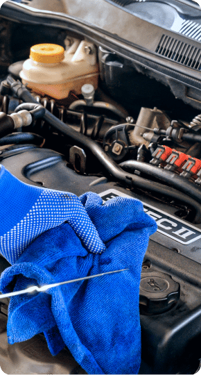 Close-up of a person wearing a blue glove and using a blue cloth to check the oil level with a dipstick under the hood of a car. The engine and various components are visible, including a plastic fluid reservoir.