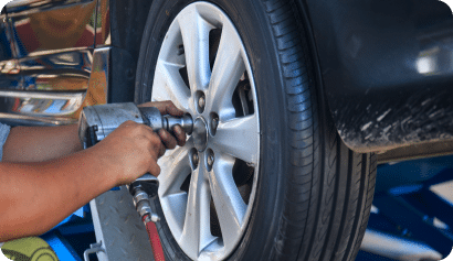 Close-up of a person using a power tool to tighten a lug nut on a car wheel. The car is elevated, and the focus is on the wheel and tire, with parts of the car's body visible in the background.