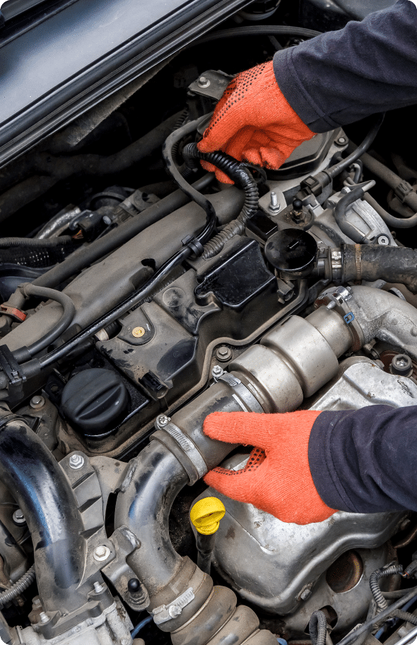 A person wearing bright orange gloves works on a car engine. The hands are adjusting a wire and gripping a component. The engine has various cables and metal parts, including a yellow cap and a turbocharger. The hood of the car is open.