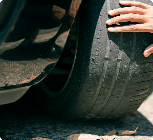 A close-up of a worn car tire with minimal tread, showing significant wear. A person's hand is inspecting the tire by running their hand along the surface, possibly checking for issues related to wheel alignment.