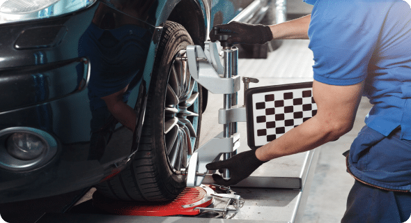 A mechanic in a blue uniform is performing a wheel alignment on a car's elevated platform. He uses a specialized tool attached to the wheel while a checkered calibration board is visible in the background, ensuring precise adjustments.