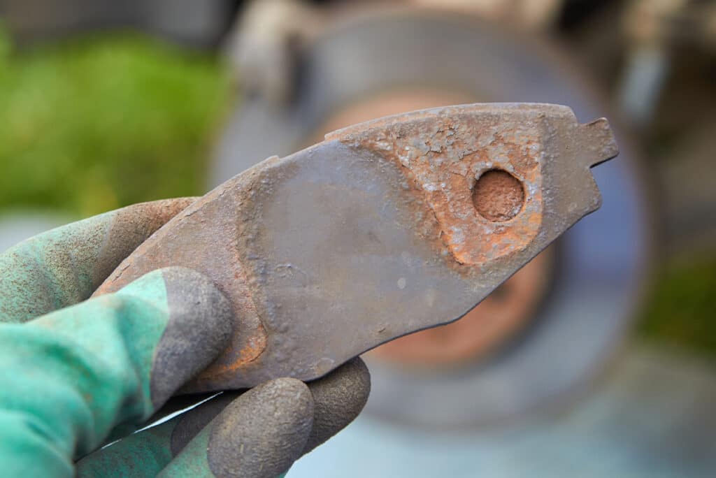 On long road trips, why do brake pads overheat on vehicles in Newport, NH with Promex Auto. Image of mechanic hand wearing green and black gloves holding up rusted and worn brake pads from car that came in for brake repair in the shop.