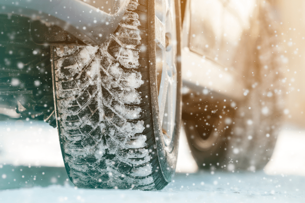 Tire repair in Newport, NH by Promex Auto. Close-up of a car tire covered in snow and ice, driving on a snowy road during winter with snowflakes falling around.